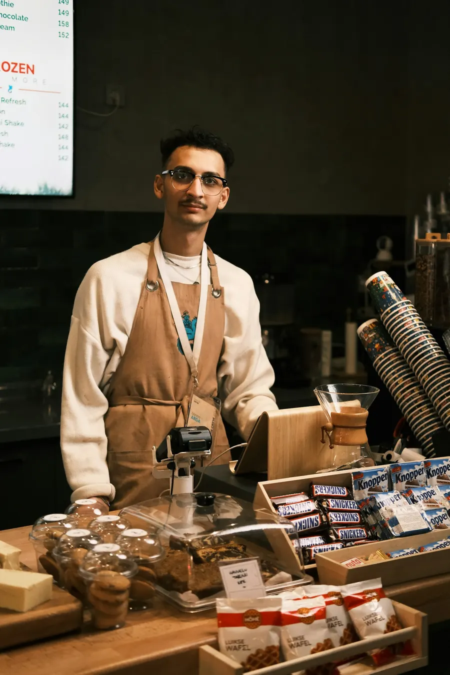 A café worker stands behind a counter of cakes and snacks